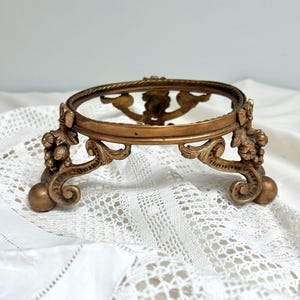 May include: A circular gold-toned metal display stand featuring ornate scrollwork, leaf motifs, and three ball-shaped feet. The stand has a raised, textured rim and an open center, showcasing intricate decorative detailing along the curved legs.
