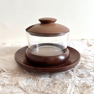 May include: A wooden lidded sugar bowl with a glass jar and a wooden base. The bowl is sitting on a white lace tablecloth.