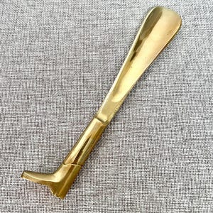 Könnte beinhalten: Ein polierter Schuhanzieher aus Messing mit einem langen, gebogenen Griff und einem kleinen, abgewinkelten Fuß. Der Schuhanzieher hat eine warme Goldfarbe und scheint aus massivem Metall zu bestehen. Das Design ist einfach und funktional.