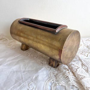 Op de afbeelding: Een cilindrische messing plantenbak met een rechthoekige opening en een donker interieur. De plantenbak staat op vier kleine, vierkante voetjes. Het messing heeft een warme, verouderde patina en de plantenbak staat op een wit kanten tafelkleed.
