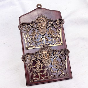 May include: A vintage wall-mounted letter rack with two compartments. The rack has a dark wood backing and decorative brass-coloured metalwork featuring a classical face design and floral patterns. A small loop is at the top for hanging.