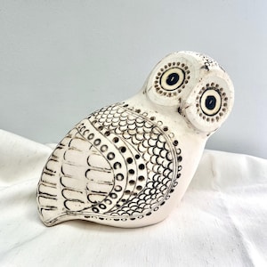May include: A cream-colored ceramic owl figurine with black detailing. The owl has large, round eyes and intricate patterns on its wings and body. The figurine is sitting on a white surface.