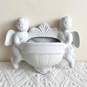May include: White ceramic wall-mounted planter featuring two cherubic figures with wings. The planter has a decorative, scalloped back and a central basin. The overall design is classical and elegant, suitable for small plants or decorative items.