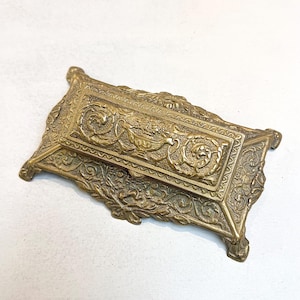 May include: Ornate brass box with a rectangular shape and intricate floral and scrollwork designs. The box has a hinged lid and small feet, showcasing a vintage aesthetic. The brass has a warm, aged patina.