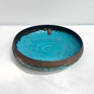May include: A small, round bowl with a bright turquoise interior and a dark bronze-coloured rim. The bowl's interior has a textured, speckled appearance. The rim has a slightly uneven, aged look.