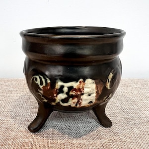 Op de afbeelding: Een donkerbruine keramische pot met drie poten. De pot heeft een brede rand en een afgeronde body met abstracte witte en bruine ontwerpen. De pot staat op een getextureerd oppervlak.