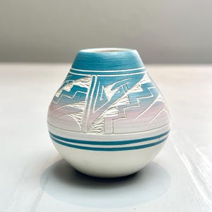 Op de afbeelding: Een kleine, handgemaakte keramische vaas met een afgeronde basis en een smalle hals. De vaas heeft een turquoise bovenkant, een witte body en een decoratieve band met geometrische patronen in roze en blauwe tinten. De vaas is ongeveer 10 cm hoog.