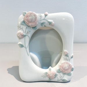 以下が含まれることがあります： 楕円形の開口部がある白い磁器製のフォトフレーム。フレームは、浮き彫りのピンクのバラと緑の葉で飾られています。フレームは、写真を飾るのに適した、角が丸い長方形の形をしています。