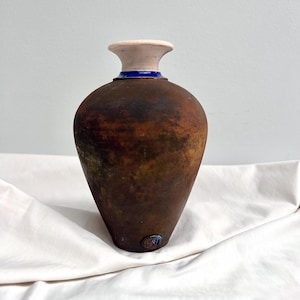 May include: A handcrafted, rustic-style vase with a brown, textured exterior. The vase features a cream-colored rim, a blue band, and a rounded body that tapers to a narrow base. The vase is likely made of ceramic or pottery.