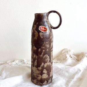 Może przedstawiać: Brązowy ceramiczny wazon z uchwytem. Wazon ma teksturowaną powierzchnię z nakrapianym wzorem brązu i beżu. Do wazonu przymocowana jest mała, okrągła naklejka z czerwonym tekstem.