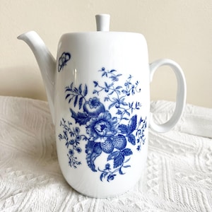 Op de afbeelding: Witte keramische theepot met een blauw bloemenpatroon. De theepot heeft een gebogen tuit, een handvat en een deksel met een kleine knop. Een blauwe vlinder staat op de zijkant. Het bloemenpatroon bevat rozen en andere bloemen.