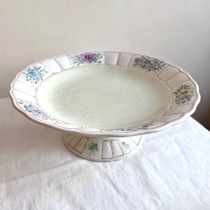 Könnte beinhalten: Ein weißer Kuchenständer aus Keramik mit einem gewellten Rand und einem Blumendesign. Der Ständer hat einen Sockel und ist mit zarten Blumenmustern in Blau-, Rosa- und Grüntönen verziert.