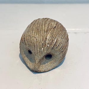 Può includere: Una scultura di gufo in ceramica con una finitura strutturata e terrosa. Il gufo ha due grandi orbite scure e un becco appuntito. La superficie è segnata da linee verticali, che le conferiscono un aspetto rustico. Un oggetto decorativo.