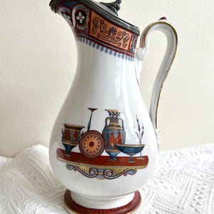 Könnte beinhalten: Eine weiße Keramikkanne mit einem Zinnverschluss und einem Henkel. Die Kanne hat ein rot-blau-goldenes Design mit einer Szene antiker griechischer Keramik.