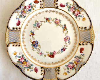 May include: A white porcelain plate with a blue and gold rim. The plate is decorated with a floral pattern in shades of pink, red, blue, and yellow.