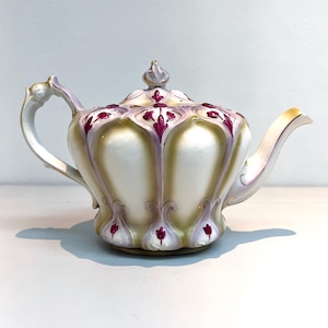 May include: A vintage porcelain teapot with a bulbous body and a lid. The teapot is primarily white with gold accents and a floral design in shades of purple and green. The handle and spout are curved.