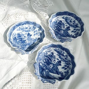 May include: Three ceramic dishes shaped like scallop shells, each featuring a blue and white transferware pattern depicting a landscape with pagodas, bridges, and willow trees. The dishes have scalloped edges and a glossy finish.