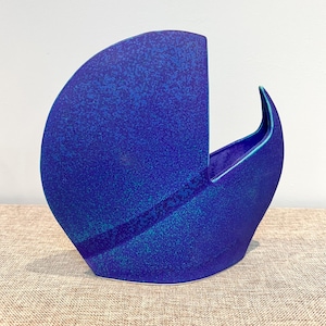 Könnte beinhalten: Eine abstrakte Keramikvase in Blautönen. Die Vase hat eine einzigartige, halbkreisförmige Form mit strukturierter Oberfläche. Das Innere ist in einem helleren Blauton gehalten, was einen Kontrast erzeugt. Ein dekorativer Artikel.