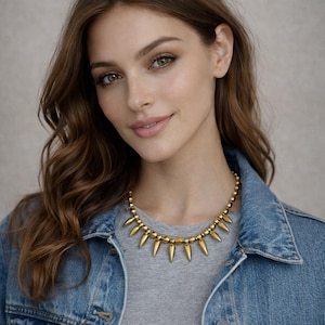 May include: A gold-colored necklace with a series of pointed, teardrop-shaped pendants. The necklace is worn over a gray t-shirt and a denim jacket. The model has long, wavy brown hair and is smiling.