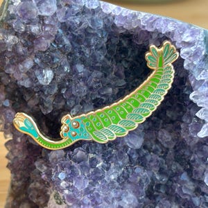 May include: A gold-toned metal enamel pin shaped like an Opabinia, an extinct marine creature. The pin features a bright green segmented body with a teal head, a long proboscis with a claw-like end, and a fan-shaped tail, all outlined in gold.