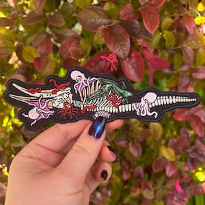 May include: Black embroidered patch featuring a skeletal crocodile, octopus, and other sea creatures. The patch has a black border and includes red, pink, and green accents. The design is detailed and intricate, suitable for jackets, bags, or other fabric items.