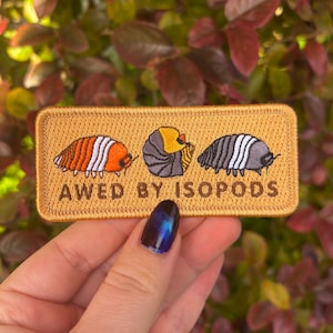 May include: A rectangular embroidered patch featuring three isopod illustrations in orange, yellow, and grey, with white detailing. The text "AWED BY ISOPODS" is below the images. The patch has a tan background and a gold border.