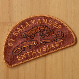 Peut inclure: Un écusson brodé marron avec le texte "#1 Salamander Enthusiast" en fil orange. L'écusson représente deux salamandres, une brune et une orange, avec des contours noirs.