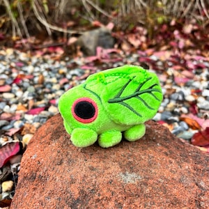 May include: A plush green caterpillar toy with a leaf-like design on its back. The caterpillar has a black eye with a red outline and small, rounded feet. A green chain is attached to the top.