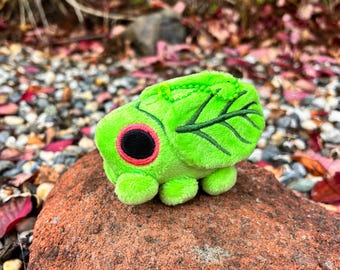 May include: A plush green caterpillar toy with a leaf-like design on its back. The caterpillar has a black eye with a red outline and small, rounded feet. A green chain is attached to the top.