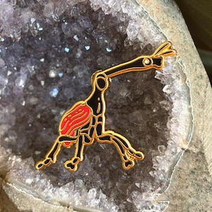 May include: A gold-colored metal pin in the shape of a stylized bird with black and red accents. The bird has a long neck and beak, with a red wing and black body. The pin is set against a background of a purple crystal.