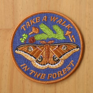 May include: Embroidered patch with a blue background and the text "Take a Walk in the Forest". The patch features a brown moth with large eyes and a green leaf with mushrooms.