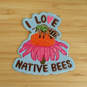 May include: An embroidered patch with a light blue background featuring a green and brown bee resting on an orange and pink coneflower. The text I LOVE NATIVE BEES is stitched in brown thread, with a small pink heart above the bee.