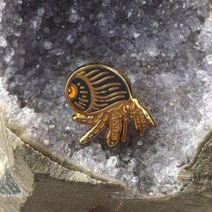 May include: A gold-colored enamel pin with a stylized design of a creature. The pin features a black body with gold accents and a small orange eye. The pin is set against a background of a purple crystal.