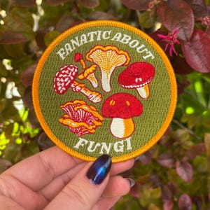 May include: Round embroidered patch with a green background and an orange border. The patch features illustrations of various mushrooms in red, yellow, and white, with the words "FANATIC ABOUT FUNGI" in white and orange lettering.