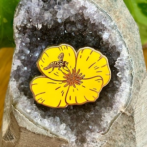 May include: A gold-tone enamel pin featuring a yellow poppy flower with a bee on it. The pin is set against a background of a purple amethyst geode.