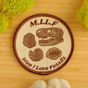 May include: Circular patch with a brown trim and a beige centre. The patch displays a T-Rex skull, a trilobite, a nautilus, and a leaf fossil. The text "M.I.L.F. Man I Love Fossils" is embroidered on the patch.