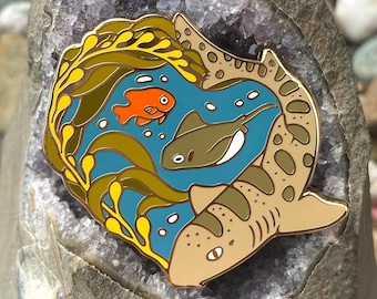 May include: Enamel pin featuring an underwater scene with a shark, fish, and stingray. The pin has a tan shark with brown spots, a blue background, and yellow and green seaweed. An orange fish and a gray stingray are also featured.