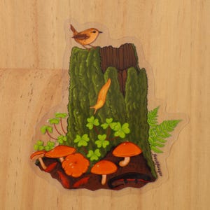 May include: A sticker featuring a whimsical illustration of a forest scene with a brown bird perched on a moss-covered tree stump, a yellow slug crawling down the stump, green clover plants, orange mushrooms, and a black and red salamander. The sticker is signed "FossilFrogger".