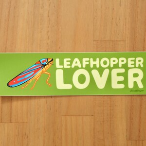 May include: A green sticker with the words "LEAFHOPPER LOVER" in large, light yellow letters. A colorful illustration of a leafhopper insect is on the left. The sticker is on a wooden surface.