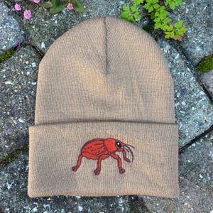 May include: Tan beanie hat with a folded brim. The hat features an embroidered red insect design on the front. The hat is made of a knitted material and is suitable for cold weather.