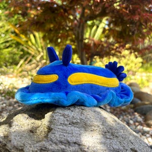 May include: A plush sea slug toy featuring a vibrant blue body with a soft, velvety texture. It has two upright antennae on top, elongated yellow patches along its sides, and a ruffled, wavy edge around the base with a cluster of blue gills.