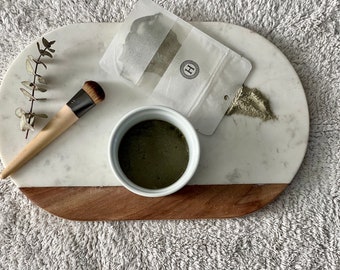 Może przedstawiać: Biała ceramiczna miska wypełniona zieloną maseczką do twarzy na marmurowej i drewnianej tacy. Obok miski znajduje się mały biały woreczek z literą "H". Na tacy znajduje się również pędzel do makijażu i gałązka eukaliptusa.