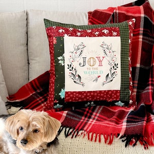 May include: A decorative pillow with a Christmas theme, featuring the text "JOY TO THE WORLD" surrounded by a wreath. The pillow is on a couch with a red and black plaid blanket. A small dog is in the foreground.