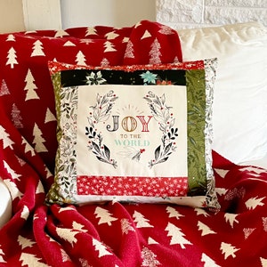 May include: Square Christmas pillow with the text "JOY TO THE WORLD" in a wreath design. The pillow has a white background with a floral wreath, and is surrounded by red, black, and green patterned fabric. A red blanket with white Christmas trees is in the background.