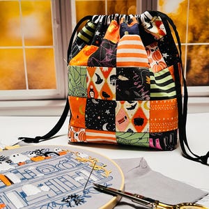 May include: A drawstring bag made of patchwork fabric featuring Halloween motifs like pumpkins, ghosts, and bats in orange, black, and green. In front, an embroidery hoop holds a fabric piece with stitched designs, alongside gold-toned scissors.
