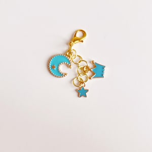 May include: A gold-colored charm with a lobster clasp, featuring a turquoise crescent moon, a star, and a crown. The moon and crown have gold outlines, and the star is a solid turquoise color. The charm is likely for a bracelet or necklace.
