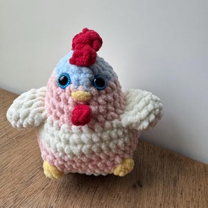 Puede incluir: Un juguete de pollo de peluche hecho a mano con cresta y barbilla rojas, ojos azules y patas amarillas. El cuerpo es rosa y blanco, con alas blancas. El juguete est&aacute; hecho de hilo suave y texturizado.
