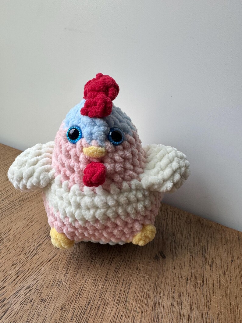 Puede incluir: Un juguete de pollo de peluche hecho a mano con cresta y barbilla rojas, ojos azules y patas amarillas. El cuerpo es rosa y blanco, con alas blancas. El juguete est&aacute; hecho de hilo suave y texturizado.