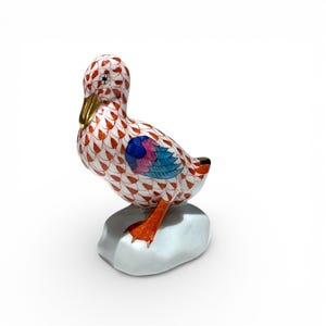 May include: A decorative ceramic duck figurine. The duck has a white body with a red and orange pattern, a gold beak, and colorful wing details. It stands on a small, light-colored base. The figurine is a collectible item.