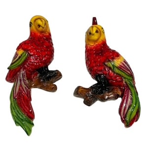 May include: Two decorative parrot figurines. The parrots are perched on brown branches and feature vibrant red, yellow, and green plumage. The figurines have a textured finish and are set against a white background.